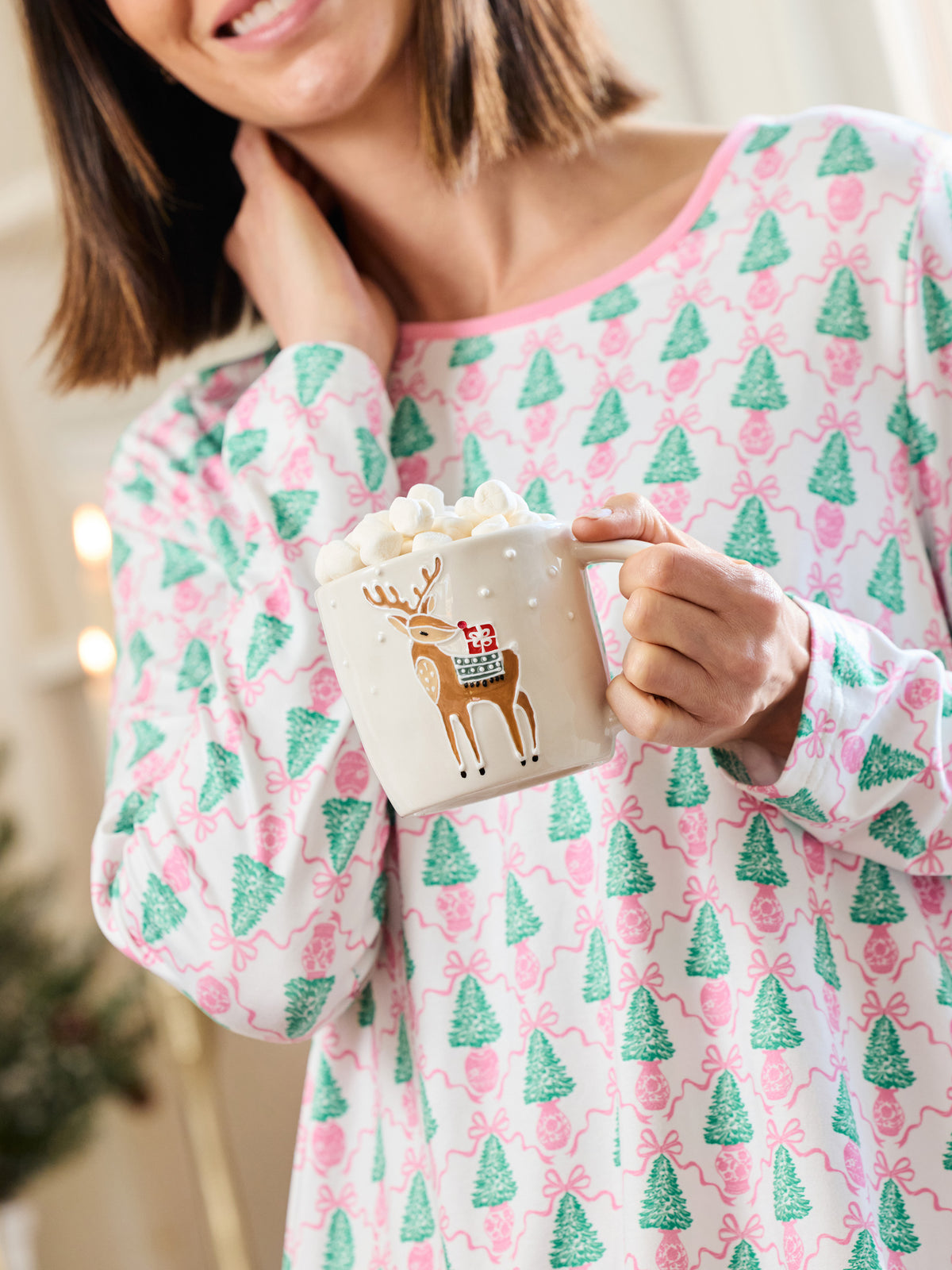 A woman wearing a pink and green holiday-themed long-sleeve pajama top holds a white mug filled with a hot drink and marshmallows. The pajama top features an all-over pattern of green Christmas trees, pink bows, and small red ornaments on a white background.