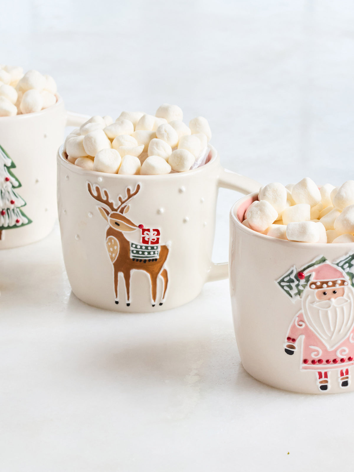 Three cream-colored ceramic mugs filled with marshmallows on a white surface. The center mug features a brown reindeer wearing a red and green sweater, and the right mug shows a pink Santa Claus.