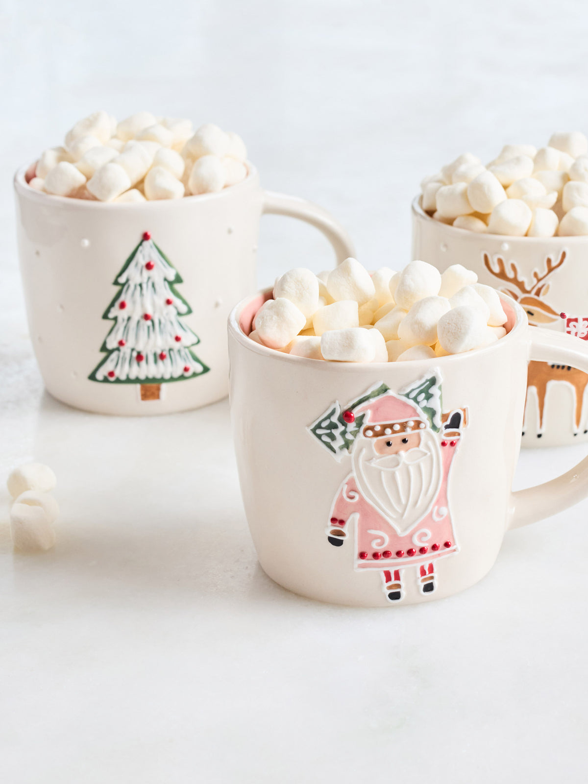 Three cream-colored ceramic mugs filled with marshmallows and hot chocolate, decorated with festive holiday illustrations including a Christmas tree, Santa Claus, and a reindeer.