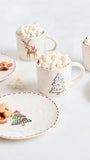 A styled holiday flat-lay featuring cream-colored ceramic mugs and plates decorated with Christmas illustrations, filled with marshmallows and topped with gingerbread cookies on a light gray surface.