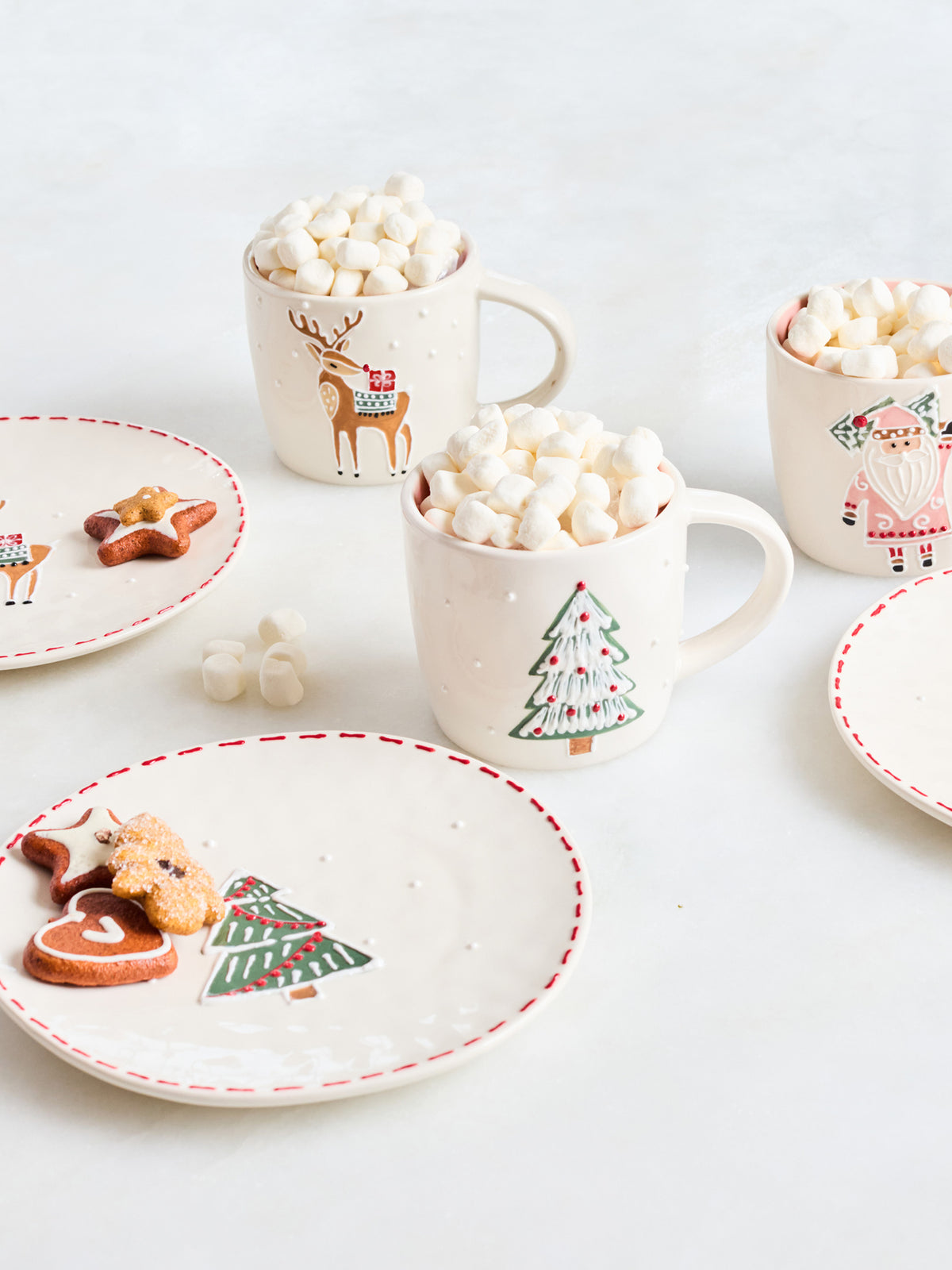 A styled holiday flat-lay featuring cream-colored ceramic mugs and plates decorated with Christmas illustrations, filled with marshmallows and topped with gingerbread cookies on a light gray surface.