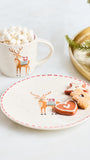 A styled holiday tabletop scene with a white ceramic mug filled with marshmallows and a matching white plate featuring a reindeer illustration and red dashed rim. A gold tray with pine branches sits in the background on a light surface.
