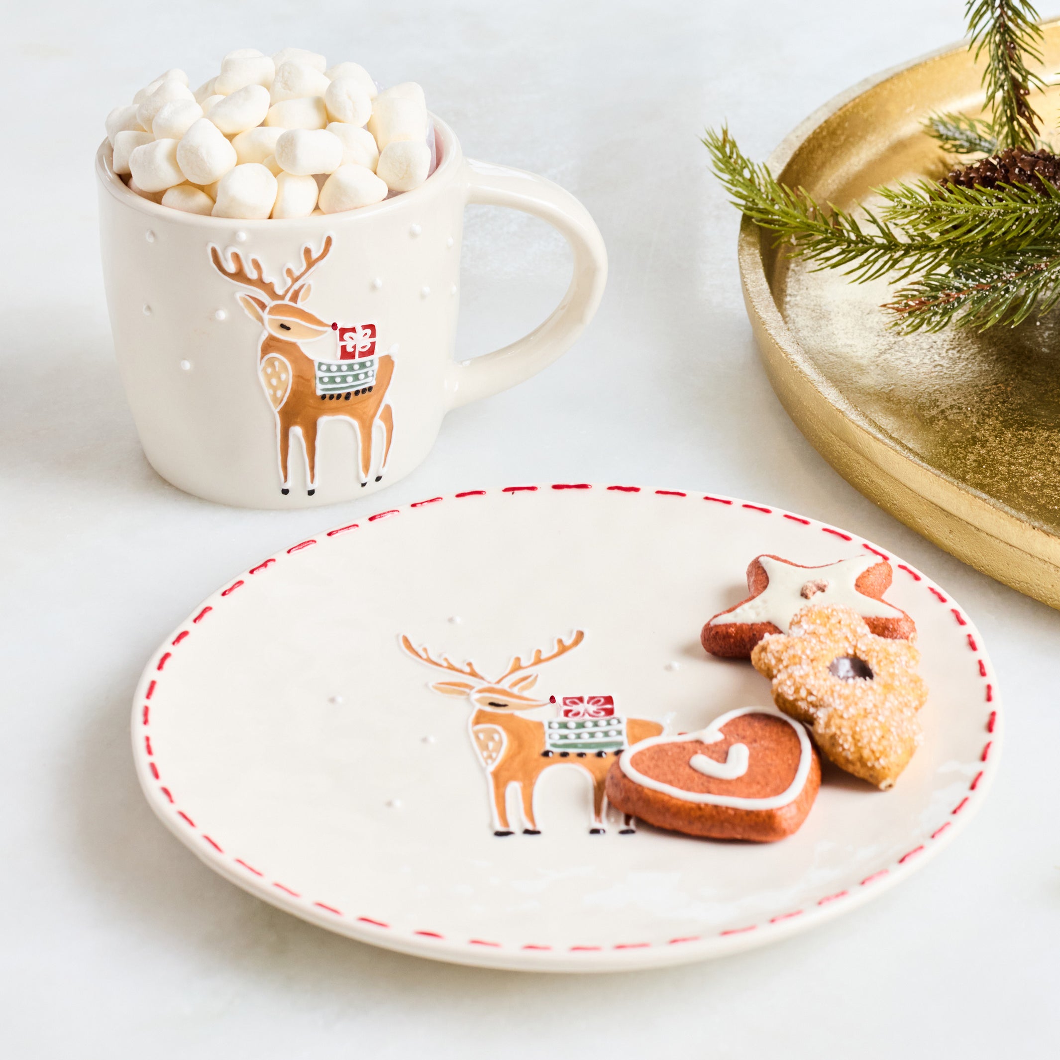 A styled holiday tabletop scene with a white ceramic mug filled with marshmallows and a matching white plate featuring a reindeer illustration and red dashed rim. A gold tray with pine branches sits in the background on a light surface.