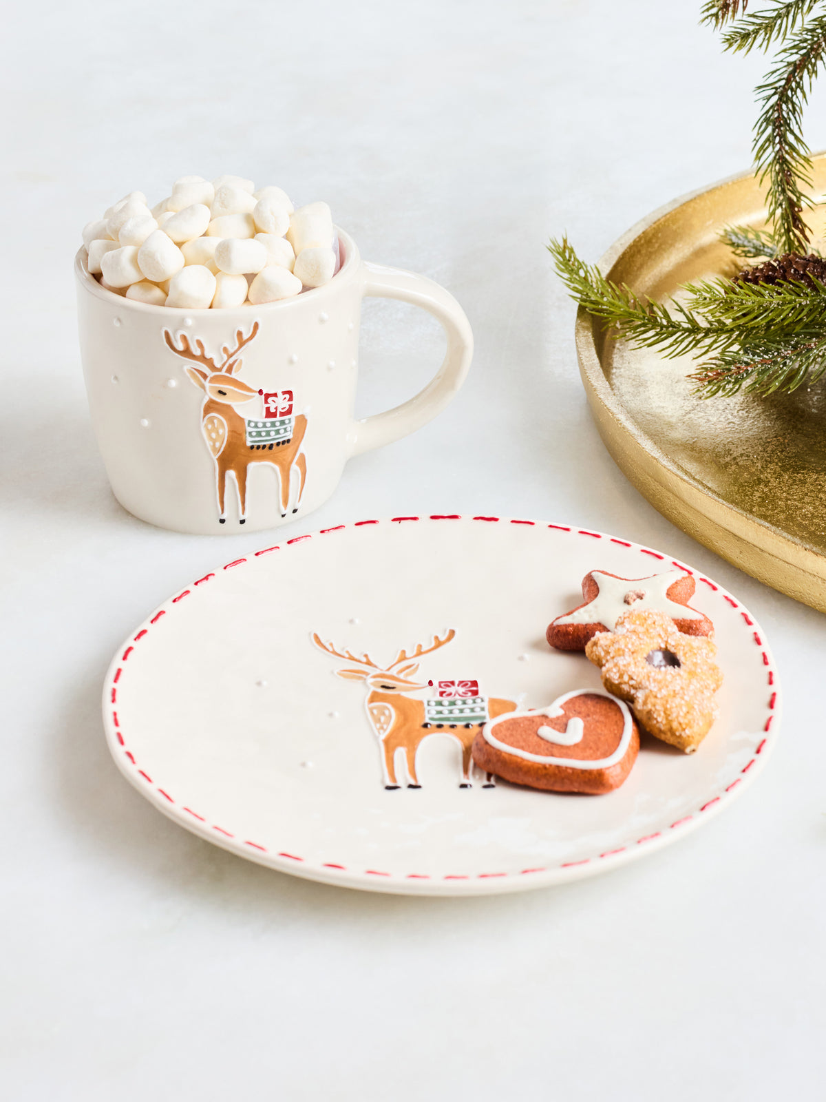 A styled holiday tabletop scene with a white ceramic mug filled with marshmallows and a matching white plate featuring a reindeer illustration and red dashed rim. A gold tray with pine branches sits in the background on a light surface.
