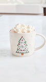 A cream-colored ceramic mug with a green and red Christmas tree design, filled with marshmallows and hot chocolate on a white marble countertop.