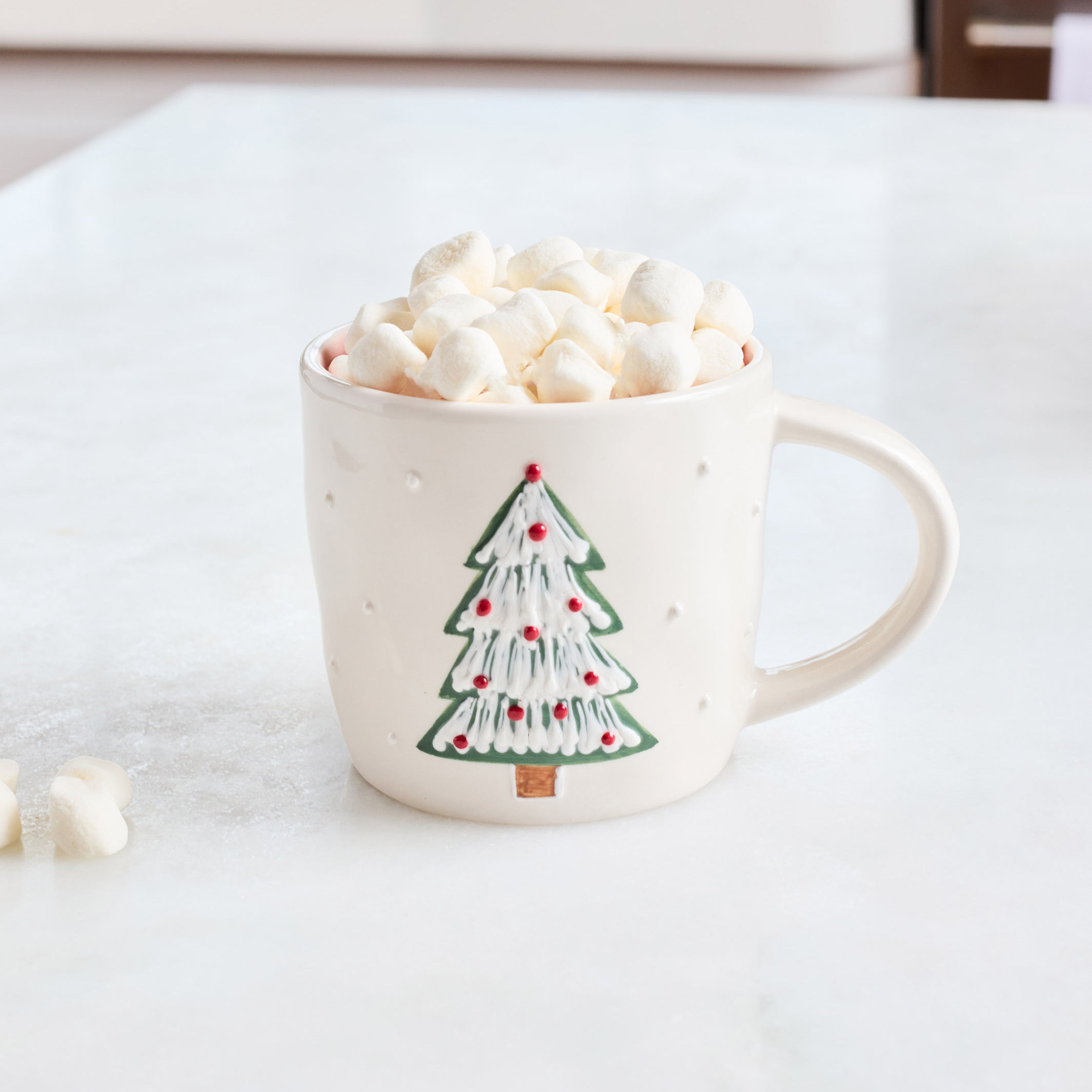 A cream-colored ceramic mug with a green and red Christmas tree design, filled with marshmallows and hot chocolate on a white marble countertop.