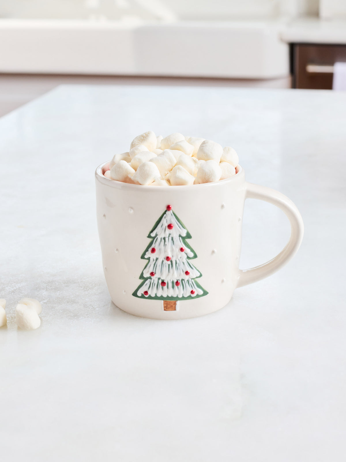 A cream-colored ceramic mug with a green and red Christmas tree design, filled with marshmallows and hot chocolate on a white marble countertop.