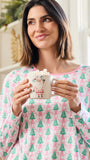 A woman wearing a pink and green Christmas-themed long-sleeve pajama top holds a white mug with a Santa Claus illustration. The background shows a softly blurred indoor setting with warm lighting.
