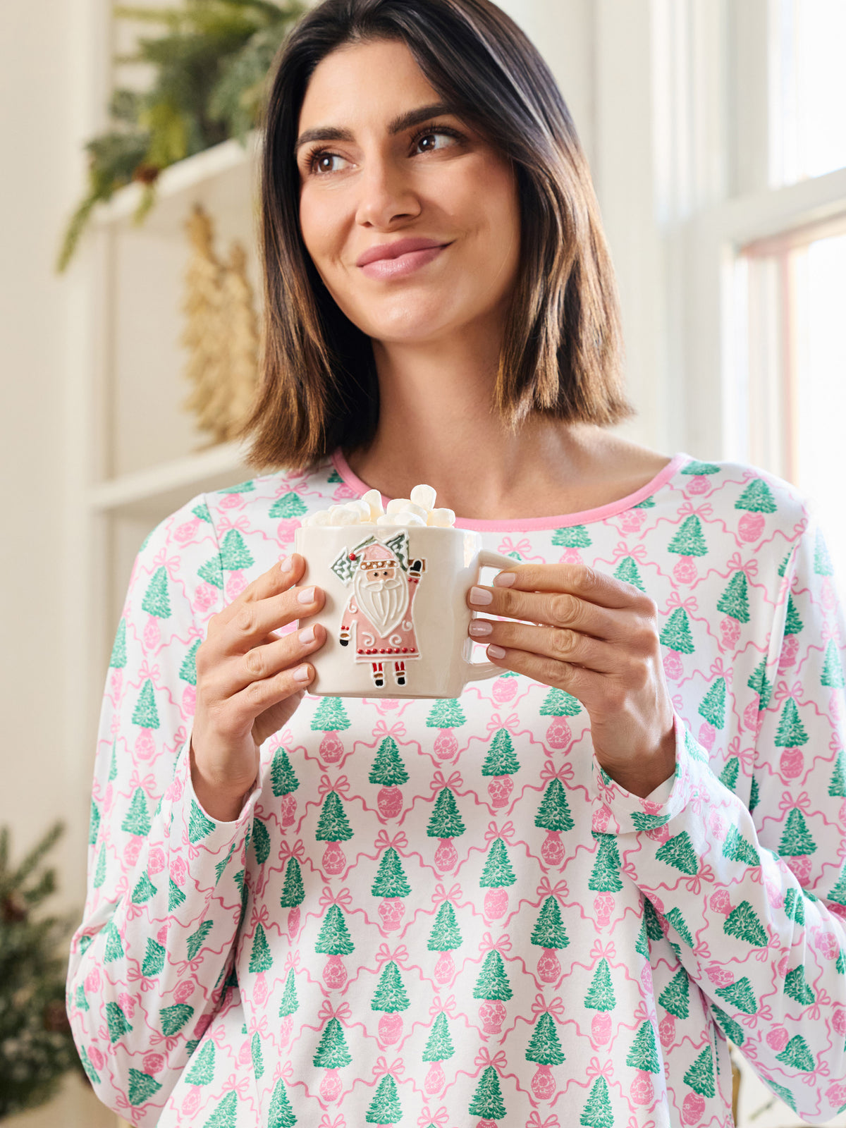 A woman wearing a pink and green Christmas-themed long-sleeve pajama top holds a white mug with a Santa Claus illustration. The background shows a softly blurred indoor setting with warm lighting.