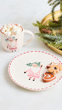 A festive holiday table setting featuring a white ceramic plate with a red dashed rim and a painted gnome, alongside a white mug filled with marshmallows and a gold tray holding evergreen branches and cookies.