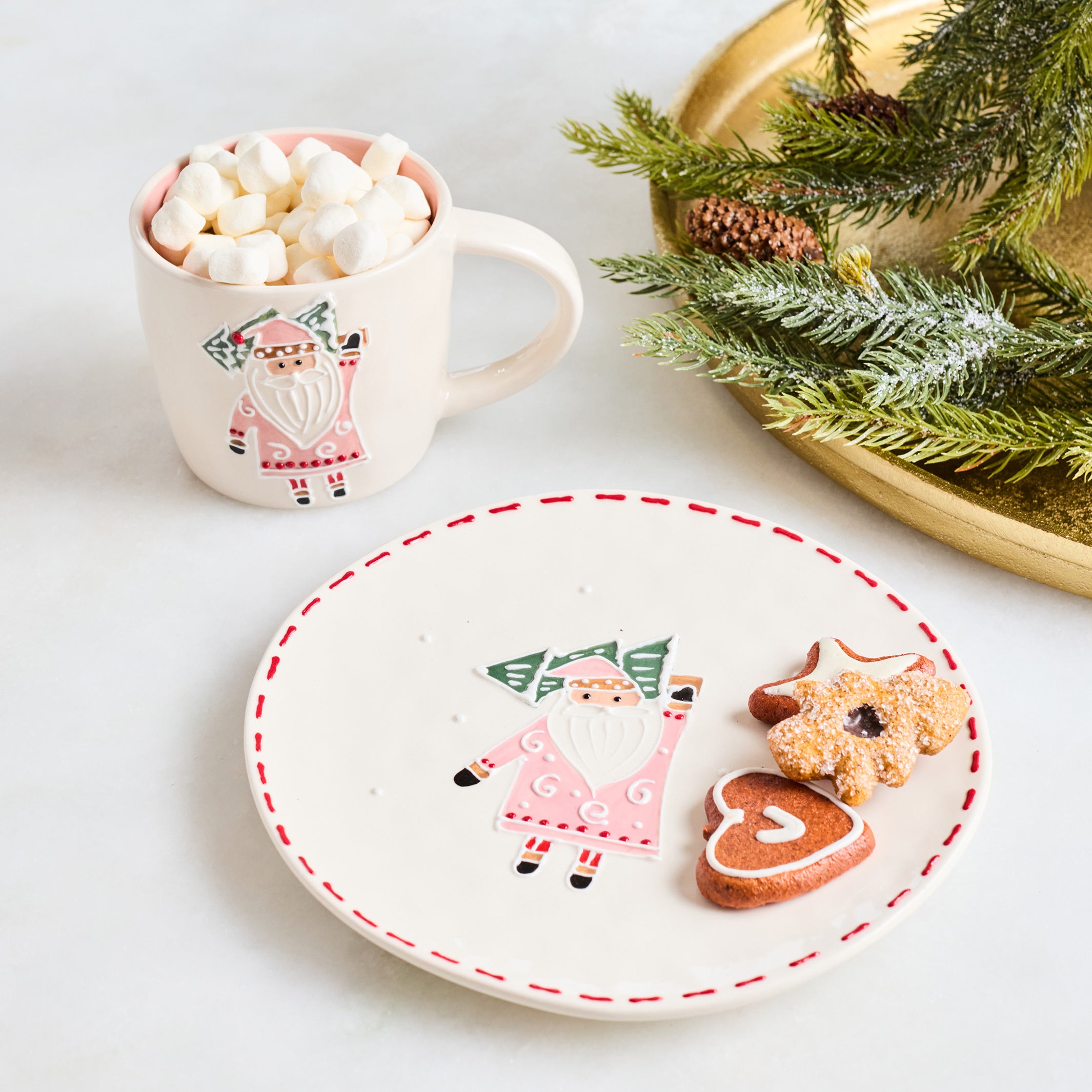 A festive holiday table setting featuring a white ceramic plate with a red dashed rim and a painted gnome, alongside a white mug filled with marshmallows and a gold tray holding evergreen branches and cookies.