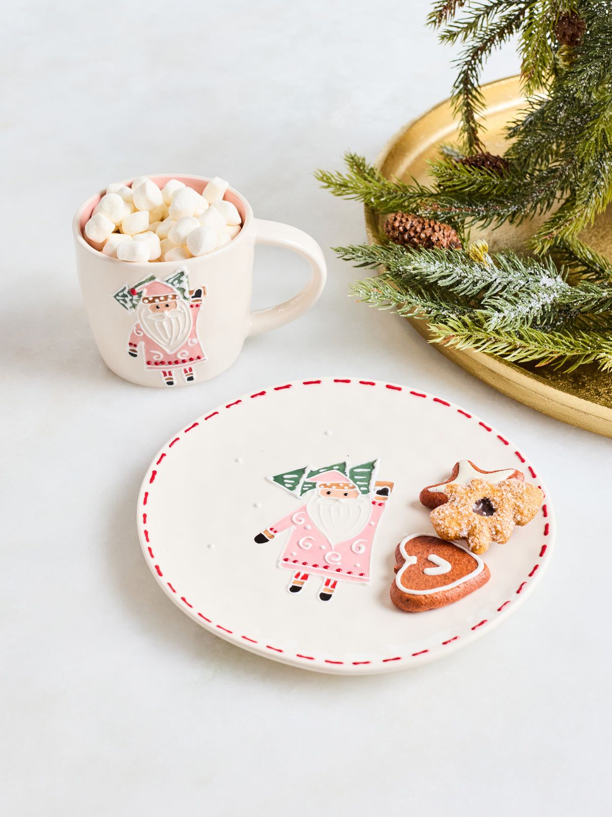 A festive holiday table setting featuring a white ceramic plate with a red dashed rim and a painted gnome, alongside a white mug filled with marshmallows and a gold tray holding evergreen branches and cookies.