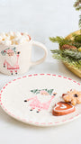 A festive holiday-themed tabletop scene featuring a white ceramic mug filled with marshmallows and hot chocolate, alongside a matching white plate with a Santa Claus illustration and red dashed rim, styled with gingerbread cookies and evergreen branches on a light surface.