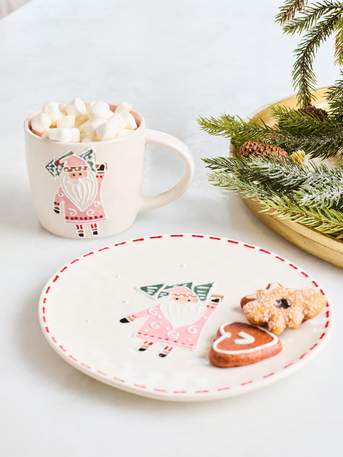 A festive holiday-themed tabletop scene featuring a white ceramic mug filled with marshmallows and hot chocolate, alongside a matching white plate with a Santa Claus illustration and red dashed rim, styled with gingerbread cookies and evergreen branches on a light surface.