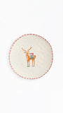 A round off-white ceramic plate featuring a central illustration of a brown reindeer with a colorful holiday sweater and the word 'MERRY'. The plate has a red dashed border and is photographed against a clean white background.