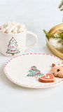 A festive holiday-themed product shot featuring a white ceramic mug filled with marshmallows and a matching white plate with a red dashed rim, both decorated with a green Christmas tree motif. Two sugar-dusted gingerbread cookies shaped like a reindeer and a heart are placed on the plate, with a gold tray and pine branch in the background.