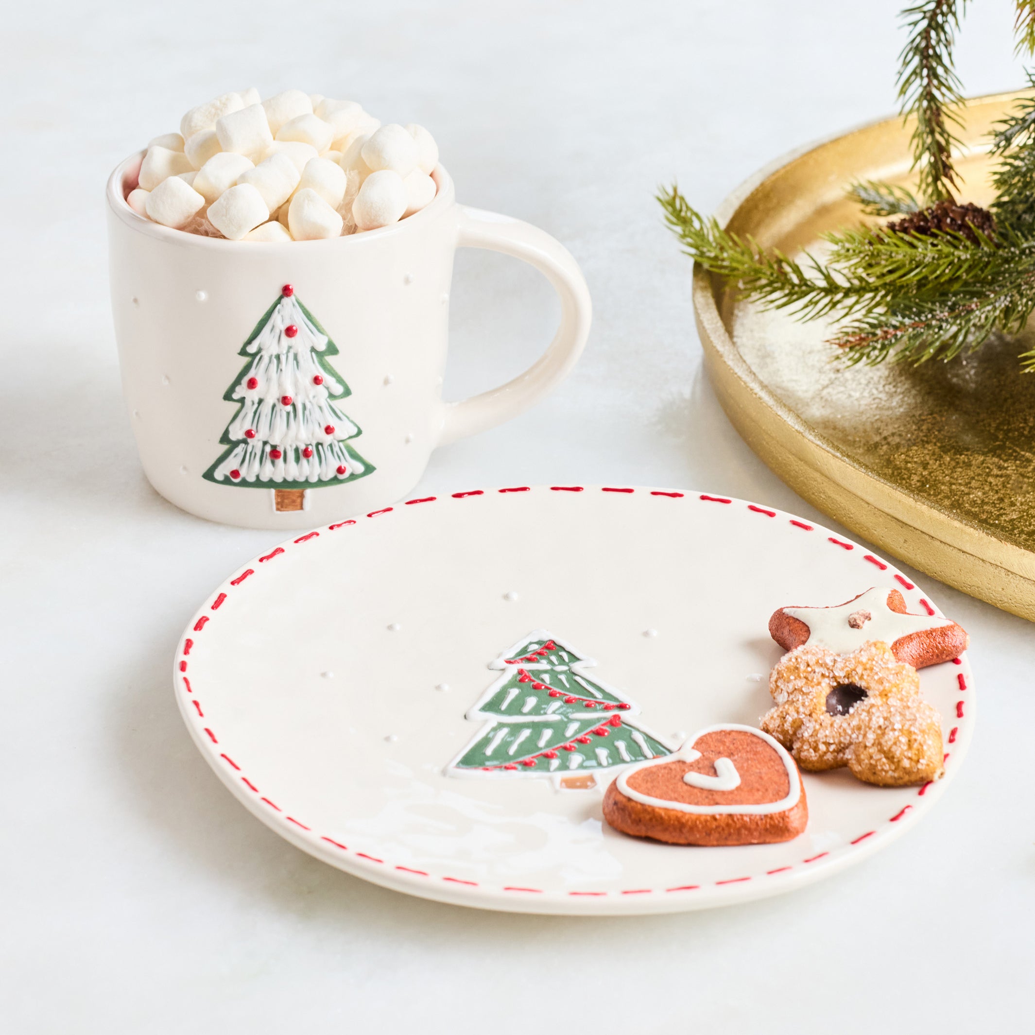 A festive holiday-themed product shot featuring a white ceramic mug filled with marshmallows and a matching white plate with a red dashed rim, both decorated with a green Christmas tree motif. Two sugar-dusted gingerbread cookies shaped like a reindeer and a heart are placed on the plate, with a gold tray and pine branch in the background.