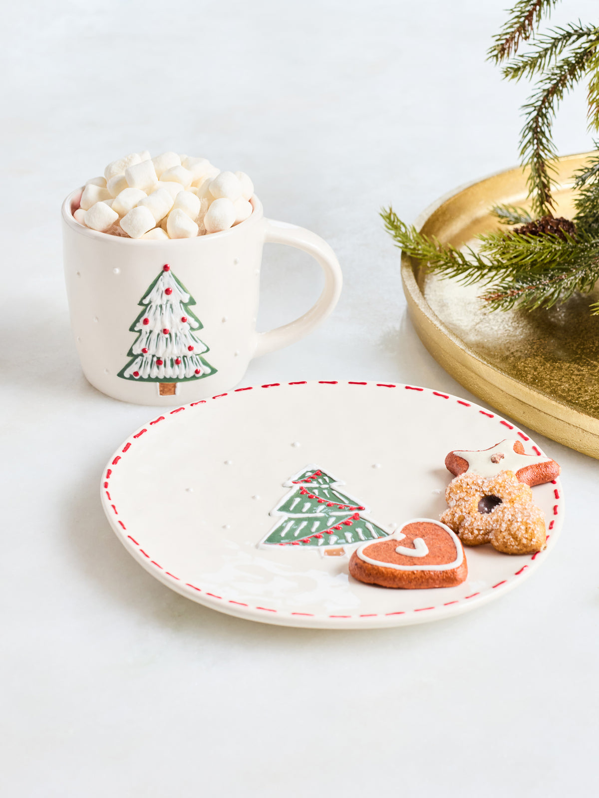 A festive holiday-themed product shot featuring a white ceramic mug filled with marshmallows and a matching white plate with a red dashed rim, both decorated with a green Christmas tree motif. Two sugar-dusted gingerbread cookies shaped like a reindeer and a heart are placed on the plate, with a gold tray and pine branch in the background.