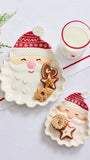 Two ceramic Christmas-themed plates shaped like Santa's face, styled with cookies and a glass of milk on a white surface with holiday greenery and a gold bell.