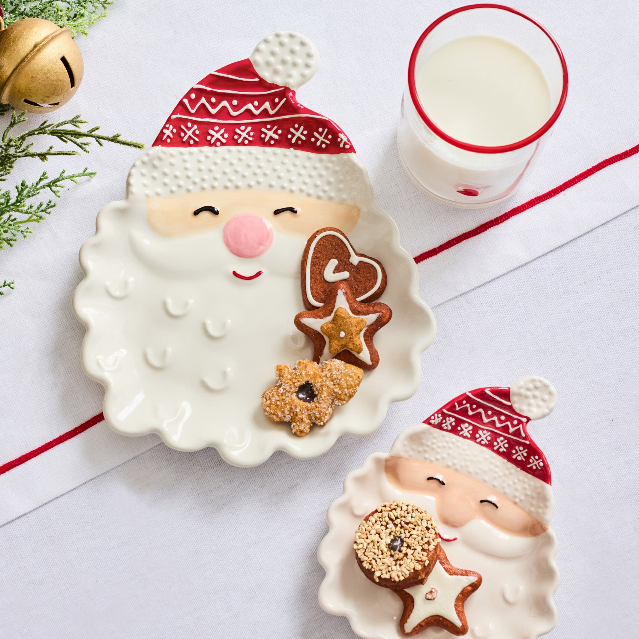 Two ceramic Christmas-themed plates shaped like Santa's face, styled with cookies and a glass of milk on a white surface with holiday greenery and a gold bell.