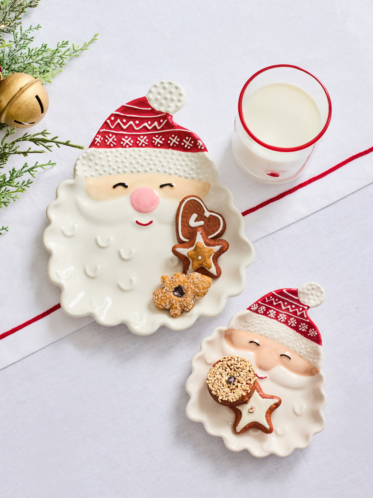 Two ceramic Christmas-themed plates shaped like Santa's face, styled with cookies and a glass of milk on a white surface with holiday greenery and a gold bell.