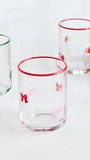 Three clear glass tumblers with festive holiday designs, including red rimmed edges and bow motifs on the front and center glasses, and wreath designs on the side glasses, set against a clean white background.