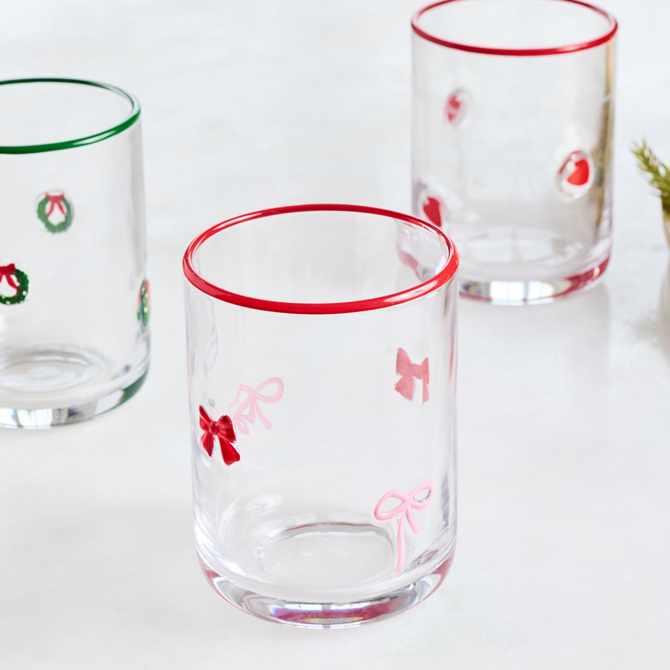 Three clear glass tumblers with festive holiday designs, including red rimmed edges and bow motifs on the front and center glasses, and wreath designs on the side glasses, set against a clean white background.