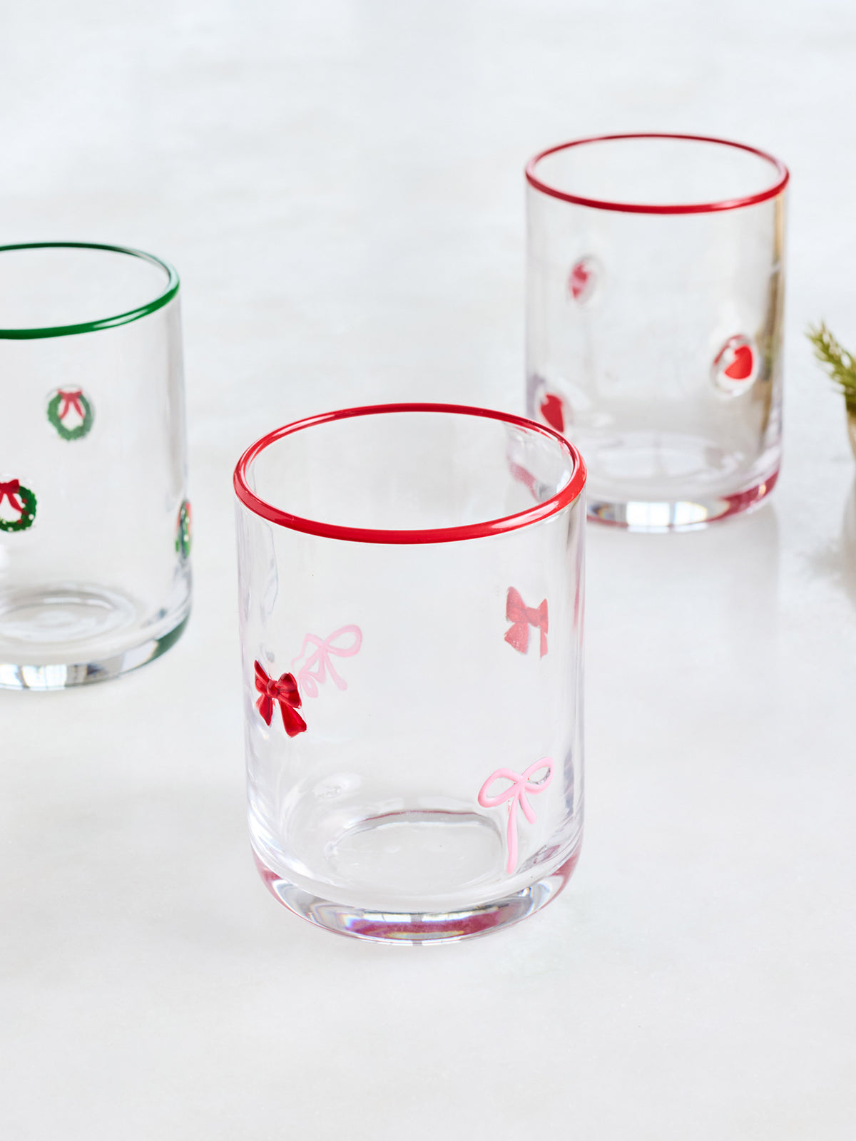 Three clear glass tumblers with festive holiday designs, including red rimmed edges and bow motifs on the front and center glasses, and wreath designs on the side glasses, set against a clean white background.