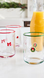 Three clear glass tumblers with holiday decorations are arranged on a white countertop next to a glass bottle of orange juice. The front tumbler has a green rim and a green wreath decoration, the left tumbler has a red rim and red bow decorations, and the back tumbler has a red rim and a small red heart.
