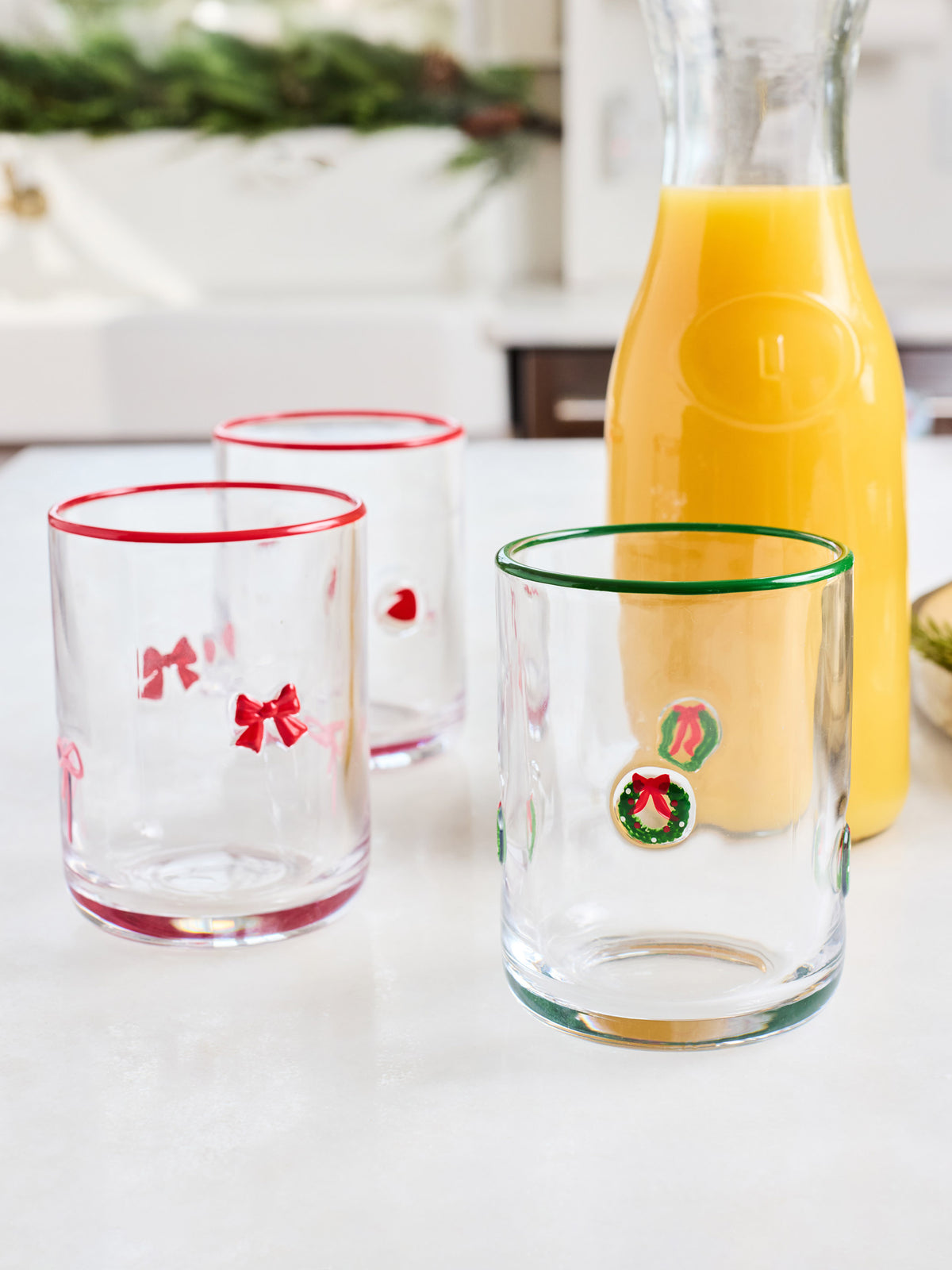 Three clear glass tumblers with holiday decorations are arranged on a white countertop next to a glass bottle of orange juice. The front tumbler has a green rim and a green wreath decoration, the left tumbler has a red rim and red bow decorations, and the back tumbler has a red rim and a small red heart.