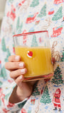 A hand holding a clear glass tumbler filled with orange juice, featuring a small red and white Santa face graphic on the side. The person is wearing a white long-sleeve pajama shirt with a colorful Christmas print of Santa Claus, trees, and sleighs.