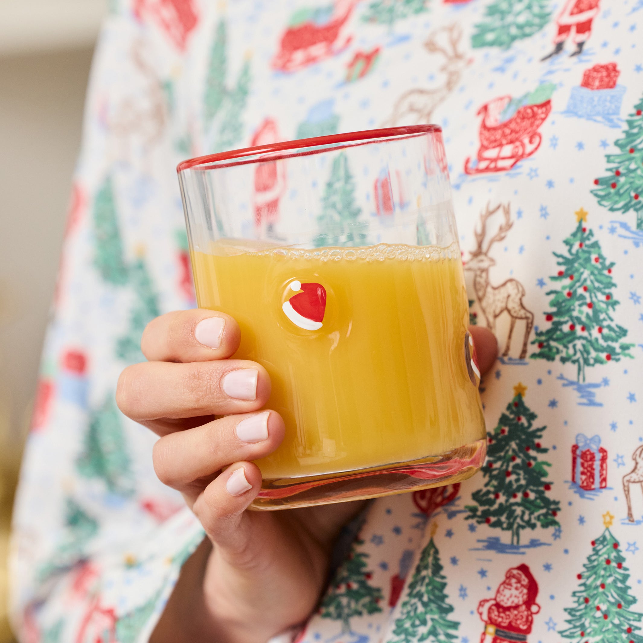 A hand holding a clear glass tumbler filled with orange juice, featuring a small red and white Santa face graphic on the side. The person is wearing a white long-sleeve pajama shirt with a colorful Christmas print of Santa Claus, trees, and sleighs.