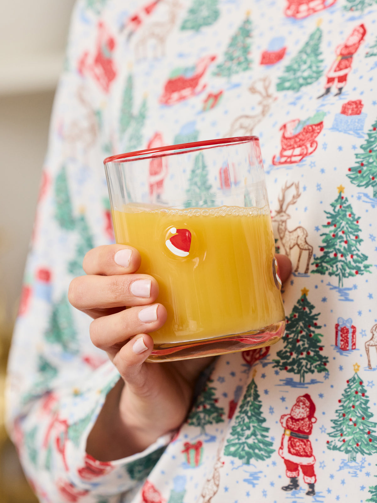 A hand holding a clear glass tumbler filled with orange juice, featuring a small red and white Santa face graphic on the side. The person is wearing a white long-sleeve pajama shirt with a colorful Christmas print of Santa Claus, trees, and sleighs.