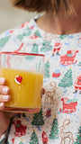 A person in a white Christmas-themed pajama shirt holds a clear glass with a red rim filled with orange juice. The pajama shirt features a festive print of Santa Claus, reindeer, and Christmas trees.