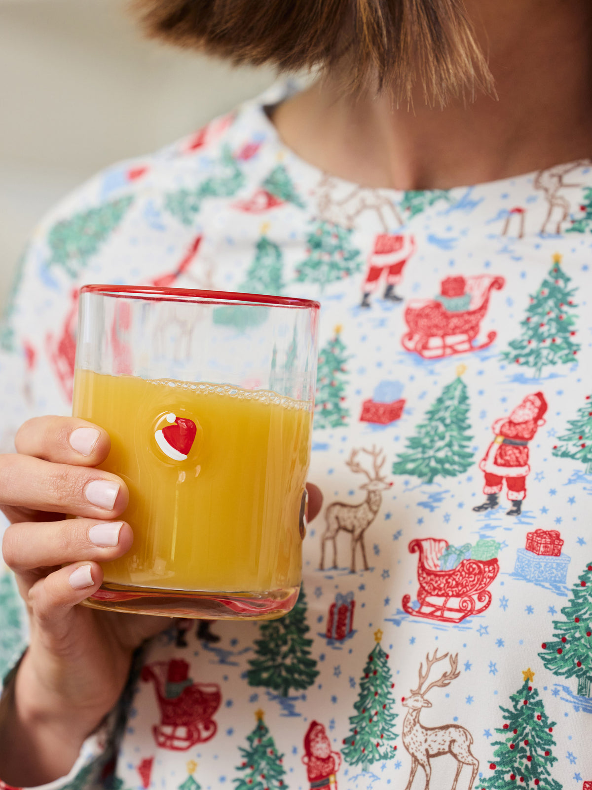 A person in a white Christmas-themed pajama shirt holds a clear glass with a red rim filled with orange juice. The pajama shirt features a festive print of Santa Claus, reindeer, and Christmas trees.