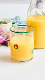 A clear glass tumbler with a green rim and small holiday wreath graphics, filled with orange juice, shown in front of a matching glass carafe on a white tabletop with food props like onion, bread, and herbs.