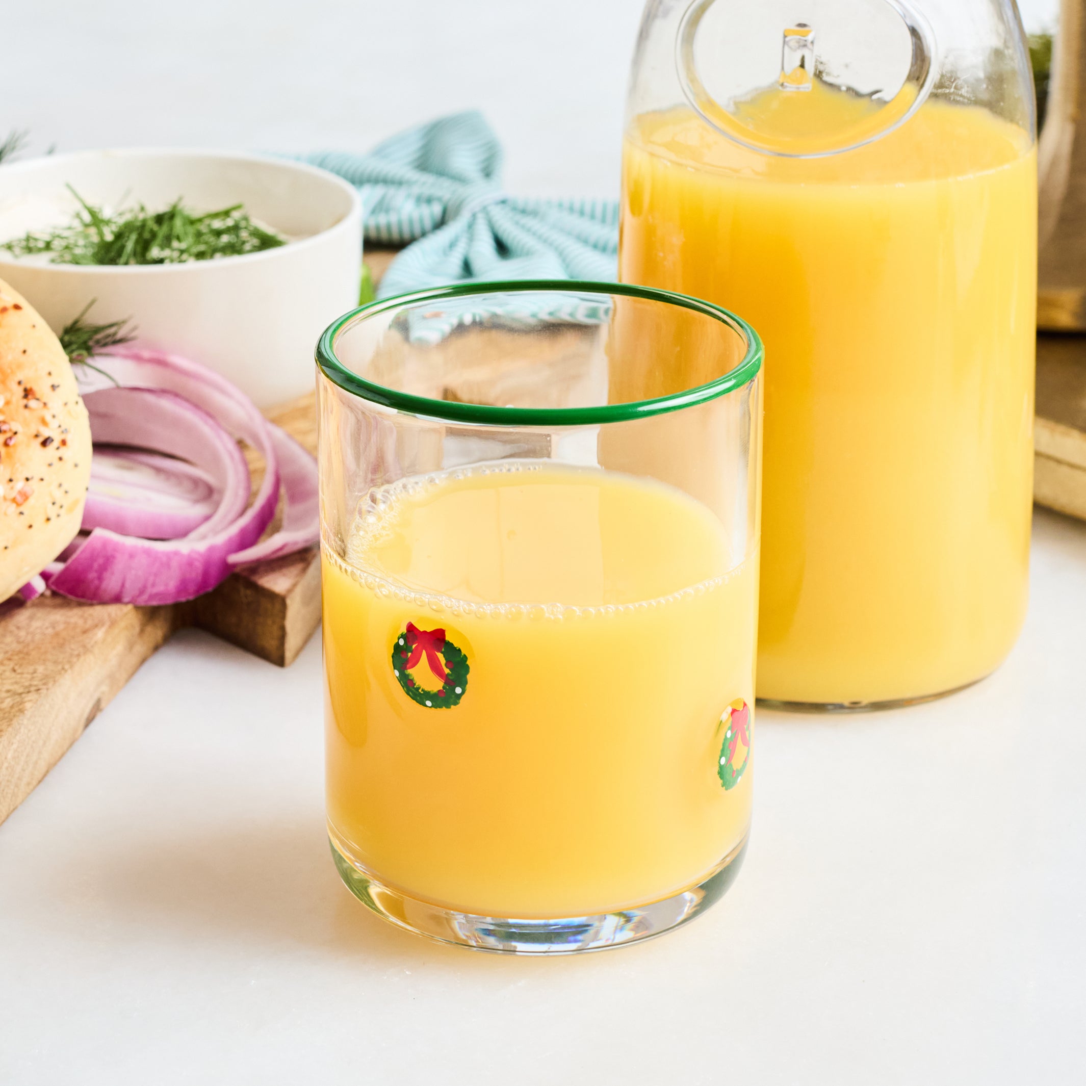 A clear glass tumbler with a green rim and small holiday wreath graphics, filled with orange juice, shown in front of a matching glass carafe on a white tabletop with food props like onion, bread, and herbs.