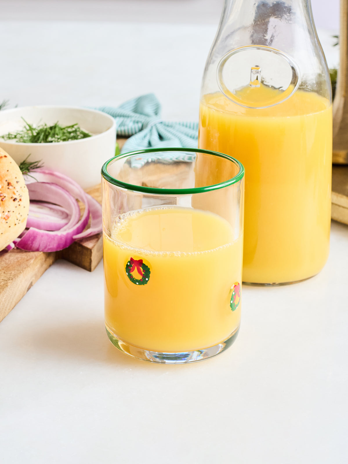 A clear glass tumbler with a green rim and small holiday wreath graphics, filled with orange juice, shown in front of a matching glass carafe on a white tabletop with food props like onion, bread, and herbs.