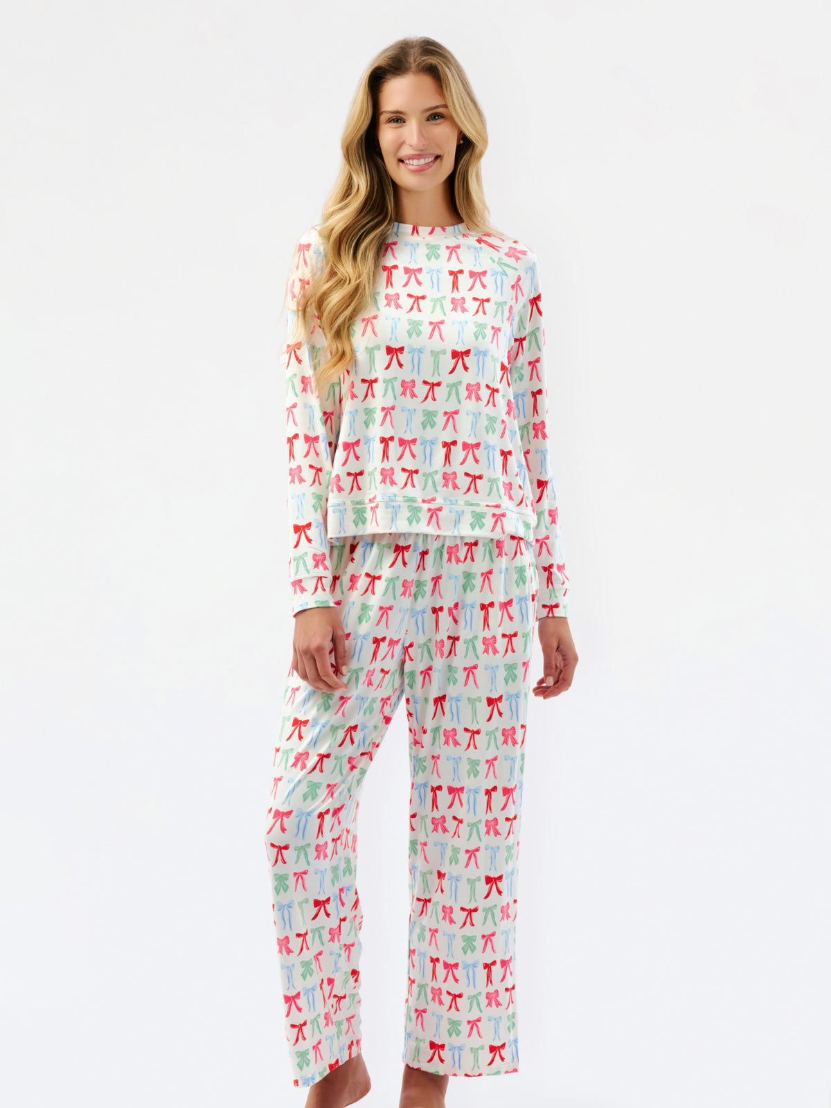 A woman modeling a matching two-piece pajama set with a long-sleeve top and wide-leg pants, featuring an all-over repeating bow pattern in red, green, and blue on a white background.