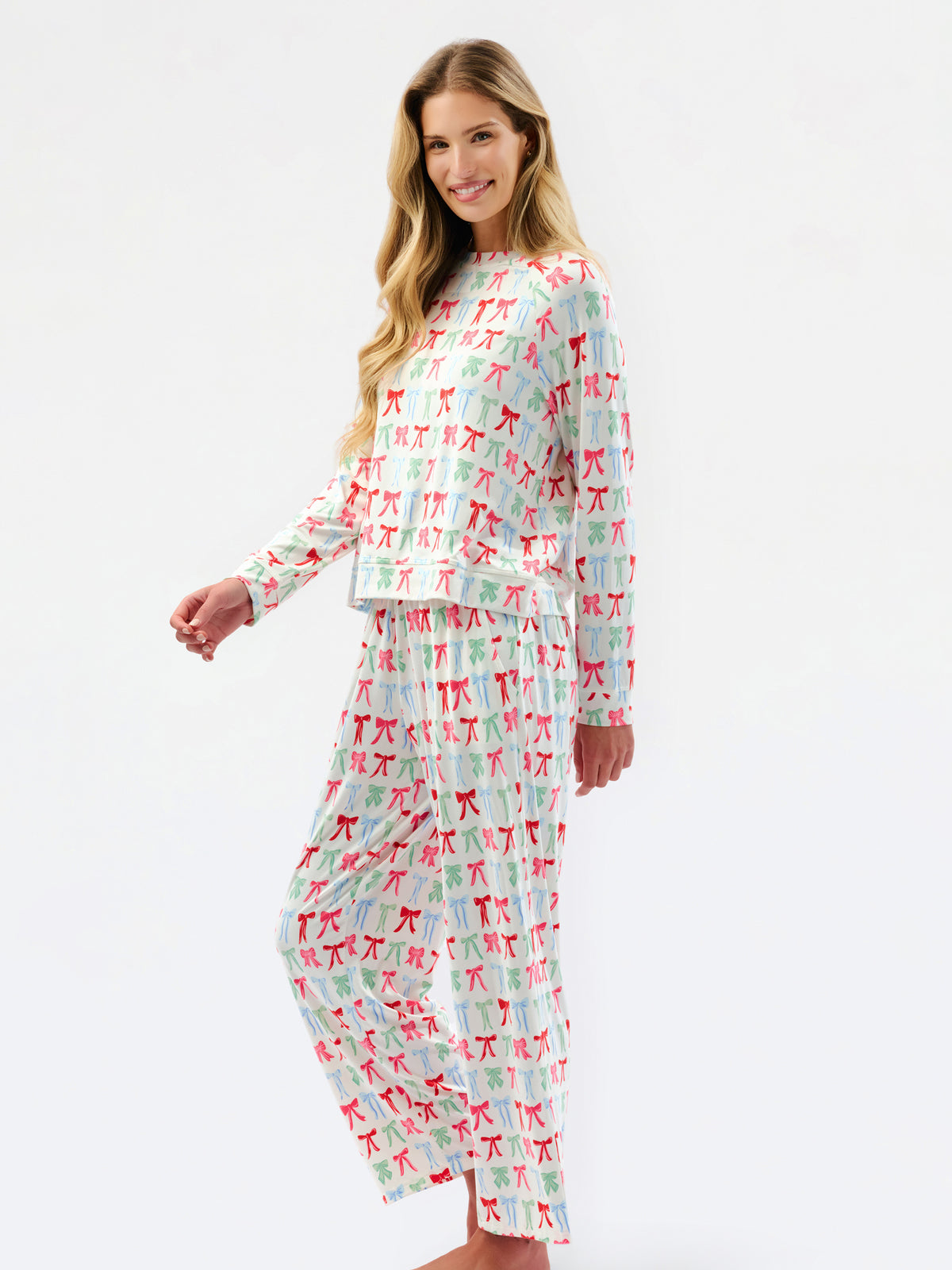 A woman modeling a matching long-sleeve pajama set with a white base and an allover print of small red and green bows, photographed against a plain white studio background.
