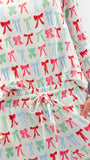 A close-up of a person wearing a matching long-sleeve pajama top and shorts set with an all-over bow print. The pajamas feature a white base with repeating red, pink, and green bows, and the shorts have a white drawstring waist.