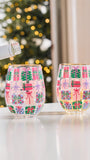 Two clear stemless wine glasses decorated with a colorful Christmas gift pattern, shown on a white table with a blurred Christmas tree in the background.