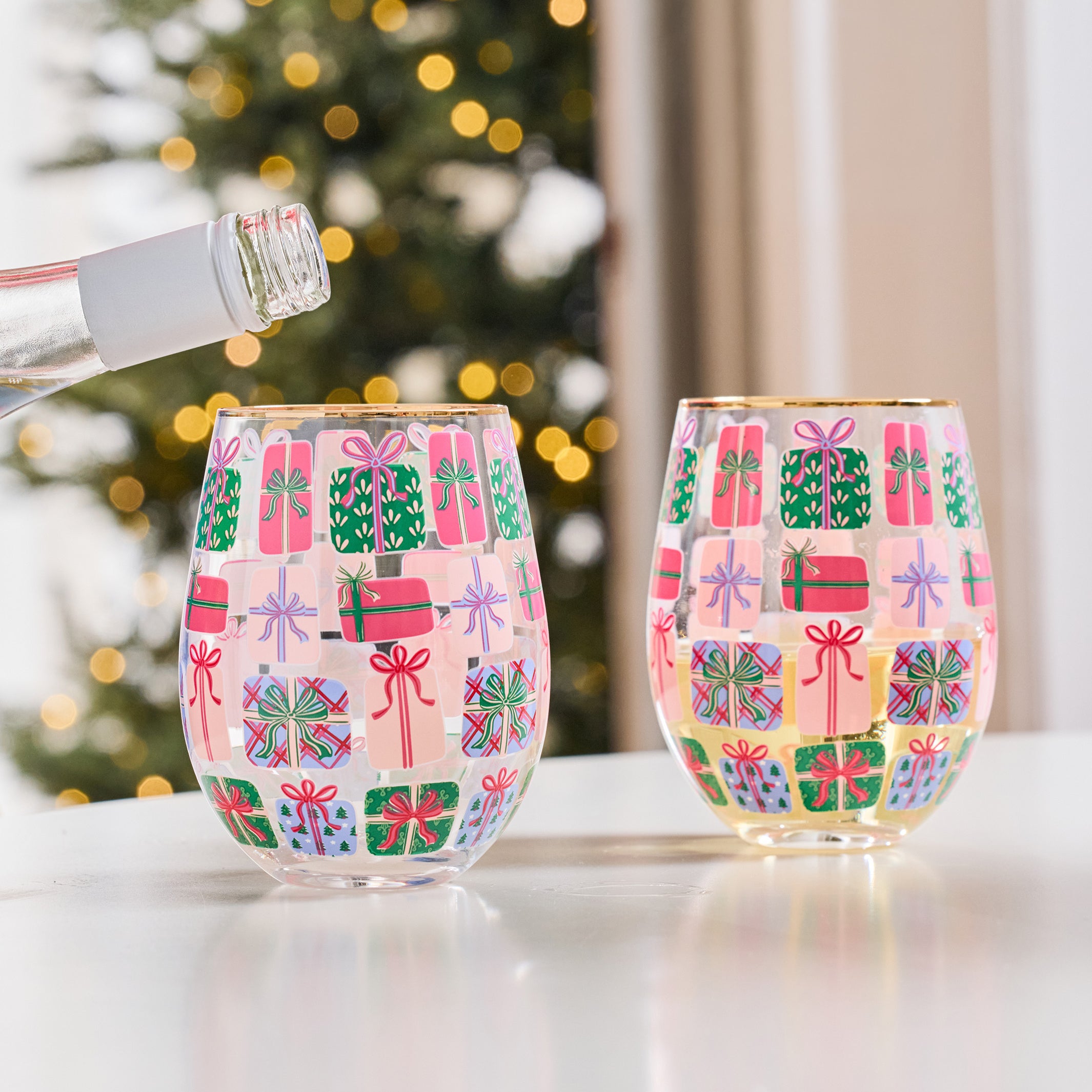 Two clear stemless wine glasses decorated with a colorful Christmas gift pattern, shown on a white table with a blurred Christmas tree in the background.