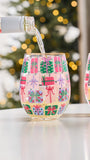 A stemless wine glass with a festive pattern of colorful gift boxes, with a bottle pouring liquid into it, set against a blurred Christmas tree background.