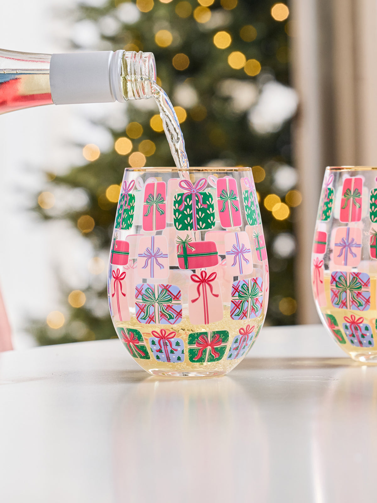 A stemless wine glass with a festive pattern of colorful gift boxes, with a bottle pouring liquid into it, set against a blurred Christmas tree background.