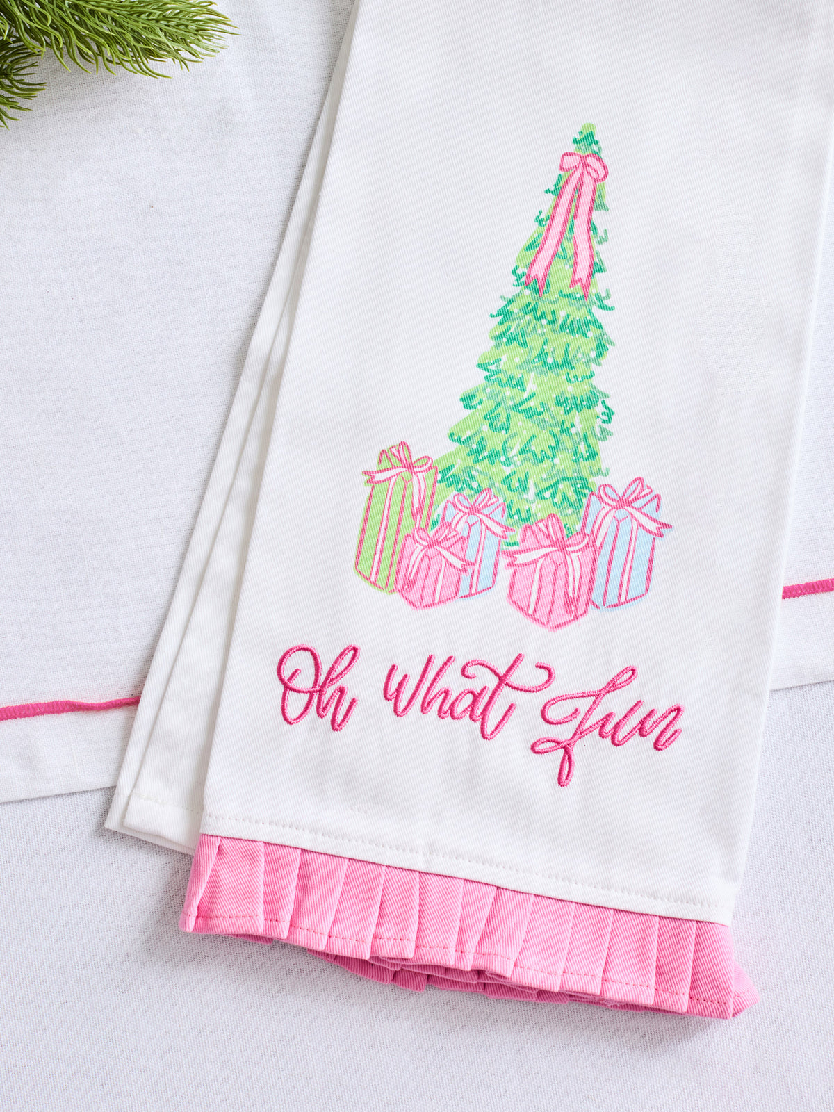 A white kitchen towel with a pink ruffled hem, featuring a colorful embroidered Christmas tree, presents, and the phrase 'Oh what fun' in pink script.