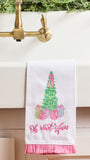 A white kitchen towel with a pink ruffled hem hangs over a white sink. The towel features an embroidered green Christmas tree with pink ribbons and wrapped presents, with the phrase 'Oh what a gem' embroidered below in pink script.