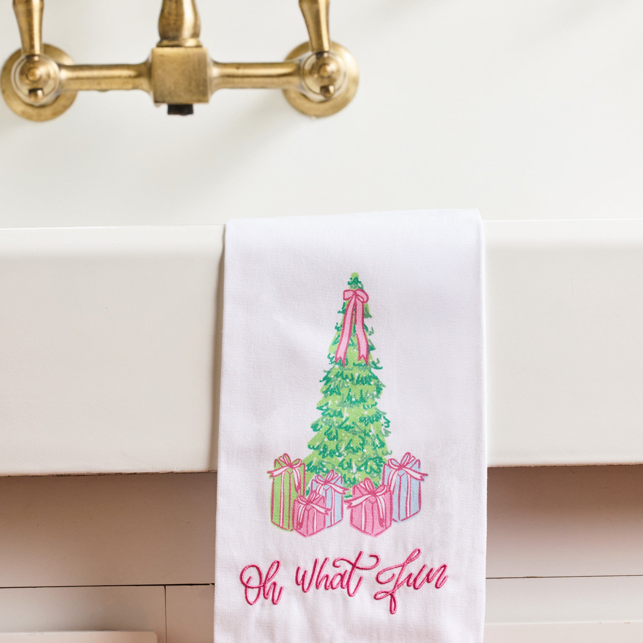 A white kitchen towel with a pink ruffled hem hangs over a white sink. The towel features an embroidered green Christmas tree with pink ribbons and wrapped presents, with the phrase 'Oh what a gem' embroidered below in pink script.