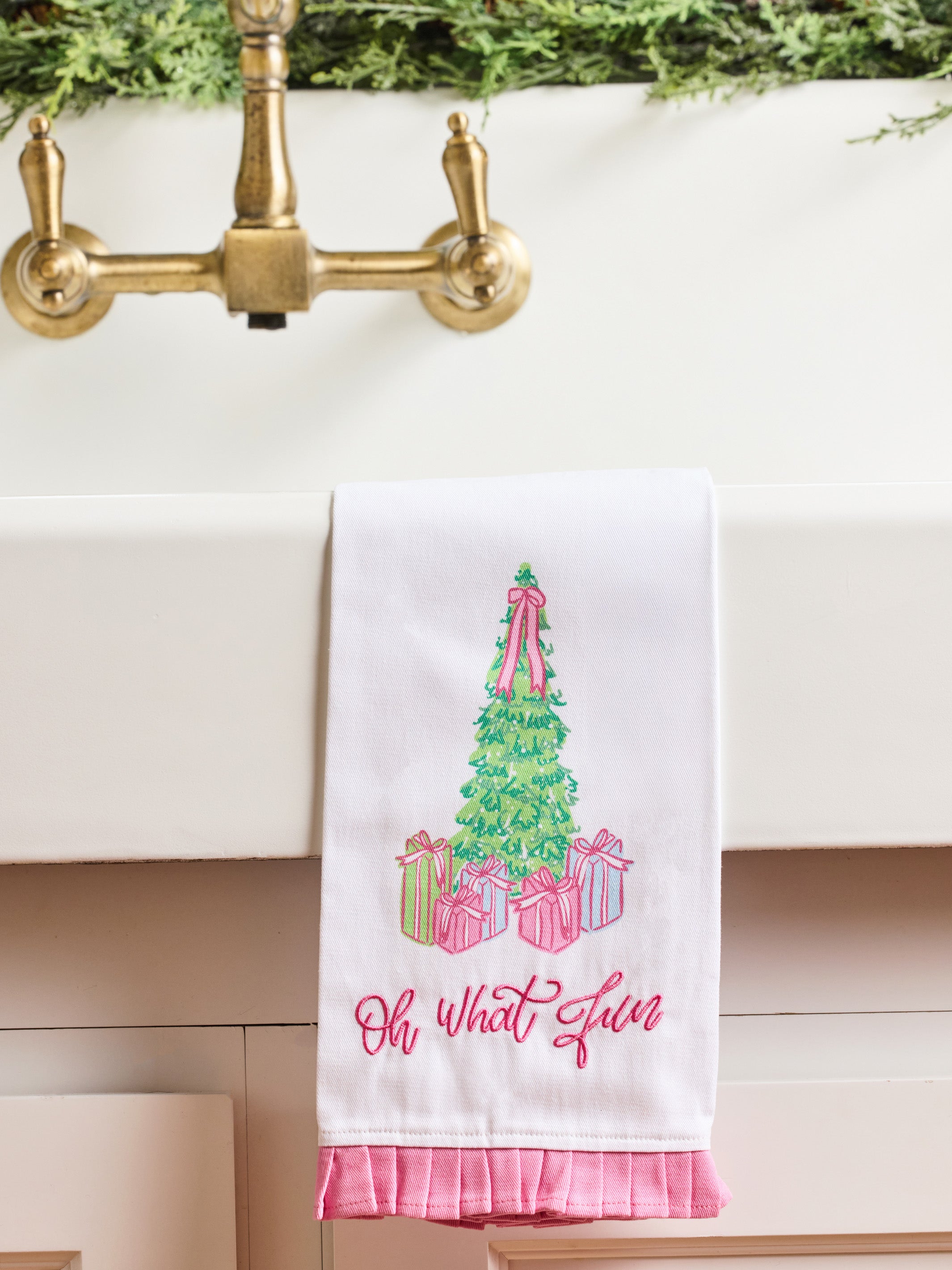 A white kitchen towel with a pink ruffled hem hangs over a white sink. The towel features an embroidered green Christmas tree with pink ribbons and wrapped presents, with the phrase 'Oh what a gem' embroidered below in pink script.