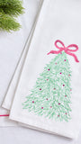 A folded white fabric napkin or tea towel featuring an embroidered green Christmas tree with a pink bow and small pink ornaments, styled on a white surface with a sprig of green pine in the corner.